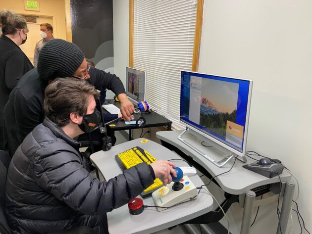 A person teaching a studeent how to use Asstive Technology. The student is sitting at a height adjustable desk, Using a large joystick and they are looking at a large screen monitor. Other equipment on this desk is a large keyboard which is yellow and black. Below the joystick is a a red button switch.