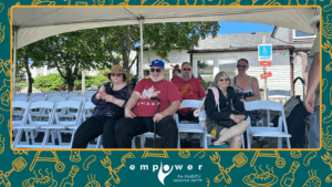 Multiple members of Empower N L sit in chairs under a large tent in front of the Centre. The Empower N L logo is centered at the bottom.
