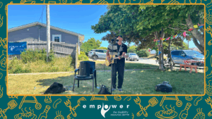 Empower N L volunteer Paddy plays guitar and sings on the front lawn. The Empower N L logo is centered at the bottom.