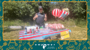 Empower N L staff person Joby runs the refreshments table at the backyard carnival. The Empower N L logo is centered at the bottom.