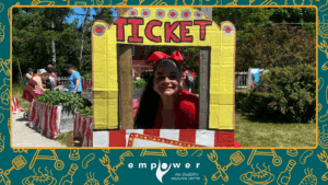 Empower N L Member Services Coordinator Sherri poses in the brightly colored carnival ticket booth. The Empower N L logo is centered at the bottom.