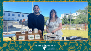 Empower N L employees Abe and Brooklyn stand behind the table of one of Empower's Youth Venture participants, selling their products. The Empower N L logo is centered at the bottom.